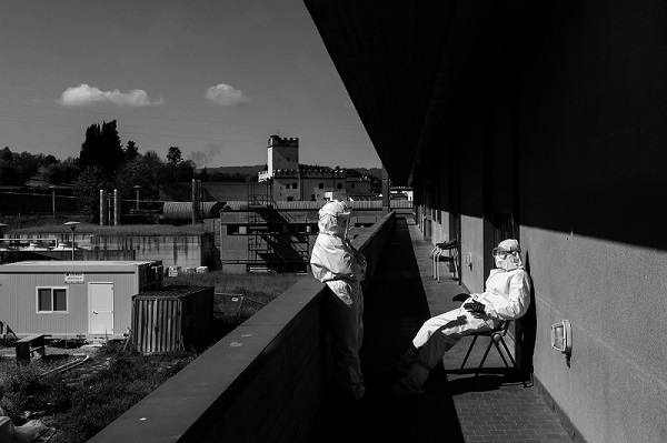 La foto è in bianco e nero, si vedono due persone con lo scafandro bianco, maschere, visiera e guanti. Una seduta su una sedia, l'altra appoggiata a un muretto, in piedi. Sono in un momento di pausa. Sullo sfondo tetti e edifici